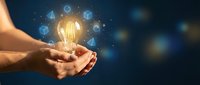 Hands holding glowing light bulb with digital innovation icons floating around on dark blue background representing creative ideas and technology solutions.