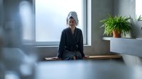 Young woman in bathrobe relaxing on wooden bench in modern spa bathroom with towel on head after shower enjoying peaceful morning wellness routine.