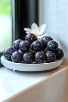 Fresh muscadine grapes on ceramic plate with white flower garnish, natural window lighting creates depth and shine on dark purple fruit skin. Food photography.
