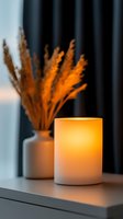 Glowing candle holder casting warm light on windowsill with dried pampas grass in white ceramic vase against dark curtain background creates cozy atmosphere.