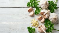 Fresh garlic bulbs and cloves with green parsley herbs arranged on rustic white wooden background for cooking and culinary preparations.