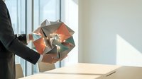 Young professional man in dark business suit holding geometric copper and gold folded paper sculpture near window with natural light, suitable for corporate design, business concepts, creativity and innovation projects.