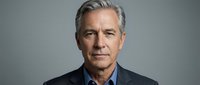 Middle-aged man with gray hair and light complexion wearing dark blazer over blue shirt, studio portrait against neutral gray background for corporate or professional use.