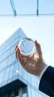 Businessman hand holding crystal sphere with heart and lightbulb icon against modern office building representing innovation and care in corporate environment.