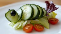 Fresh cucumber slices arranged as fish shape with olive eye on lettuce bed, garnished with cherry tomatoes, creative food art presentation for children.
