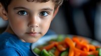 Young boy in blue shirt looking at camera with curious expression near plate of fresh colorful vegetables, focus on bright blue eyes and innocent face.