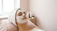 Young woman relaxing with white facial mask treatment at spa salon wearing towel on head with eyes closed for skincare wellness routine.