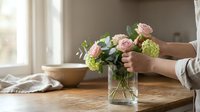 Woman arranging fresh pink roses and green flowers in glass vase on wooden table in bright home interior for decoration and lifestyle photography.