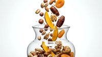 Assorted nuts and dried fruits falling into glass jar on white background, isolated for healthy snack packaging and nutrition advertising.