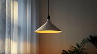 Modern pendant lamp with warm yellow light illuminating minimalist interior space with soft curtains and plant silhouette in corner.