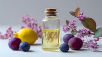 Glass bottle of essential oil with fresh plums, lemon, and pink flowers on light background, natural aromatherapy and skincare concept.