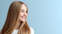 Young Caucasian woman with long straight blonde hair and natural makeup smiling against light blue background, showcasing healthy shiny hairstyle for beauty and haircare advertising.