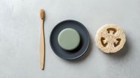 Eco-friendly bathroom essentials: bamboo toothbrush, green clay soap on dark dish, and natural loofah on light gray background.