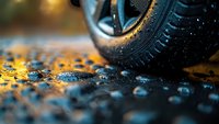 Close-up view of wet car tire on asphalt road with water droplets and golden reflections during rainy weather, creating dramatic automotive atmosphere.