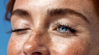 Close-up of young woman with freckles winking against blue background, showing one bright blue eye and natural lashes.