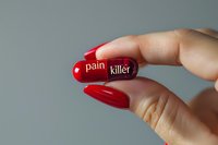 Red and purple capsule pill marked with pain killer text held between fingers with red manicure against gray background, medical and healthcare concept.