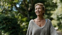 Mature Caucasian woman with short blonde hair smiling in nature, wearing a light knit sweater, representing aging gracefully and wellness.