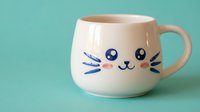 Adorable white ceramic mug with cute cat face design featuring whiskers and kawaii expression on turquoise background for kitchen decor and gifts.