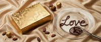 Luxury gold gift box with ornate pattern, chocolate love inscription in glass bowl with whisk, cocoa beans on silk fabric.