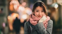 Young Asian woman wearing cozy gray sweater and pink scarf smiling warmly at camera with soft bokeh city lights background during evening hours.