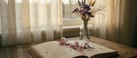 Cozy reading corner with open book, dried lavender flowers in glass vase and scattered petals on wooden table by sunlit window with soft curtains.
