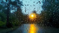 Raindrops on window glass with blurred sunset view over water, creating atmospheric golden light reflection through wet surface.