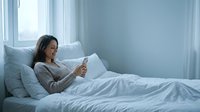 Young woman with dark hair and East Asian ethnicity relaxing in bed with smartphone, surrounded by pillows in bright bedroom with natural light from windows.