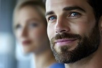 Confident bearded man with gentle smile looking forward while blonde woman stands in soft focus background. Positive emotional portrait.