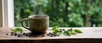 Ceramic coffee mug with fresh green leaves and coffee beans on wooden table by window. Morning beverage concept for cafes and kitchen decor.