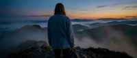 Woman with long dark hair wearing blue jacket standing on mountain peak overlooking misty valleys at sunrise, contemplating nature and landscape.