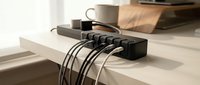 Black multi-port USB charging station organizes cables on white desk with coffee cup and books in modern minimalist workspace for electronic device management.
