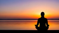 Person meditating in lotus position silhouetted against stunning sunset over calm water creating peaceful mindfulness scene for wellness and relaxation content.
