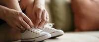Woman tying white sneakers while sitting, preparing for workout or daily activities. Close-up of hands lacing athletic shoes on wooden floor.