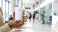Person holding white takeaway coffee cup in modern office lobby with blurred colleagues and plants in background, suitable for corporate, lifestyle, and beverage marketing.