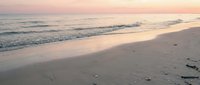 Serene beach at sunset with gentle waves washing onto sandy shore, footprints in sand, peaceful coastal scene with warm pink and orange sky colors.