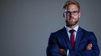 Confident male professional with light skin tone and beard wearing navy blazer, burgundy tie, and eyeglasses, arms crossed in assertive pose against neutral background for corporate portraits.