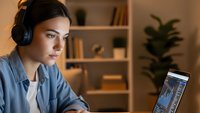 Young woman wearing headphones working on laptop computer in modern home office with bookshelf and plant in background during evening hours.