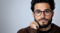 Middle Eastern man with beard and glasses wearing brown sweater poses thoughtfully against gray background, hand on chin in contemplative professional portrait.