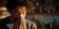 Caucasian man with curly hair blowing nose into tissue at home, suffering from cold or flu symptoms in warm evening light.