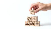 Hand placing wooden cube with palm tree icon on pyramid of travel-themed blocks featuring camera, compass, mountain and globe symbols on white background.