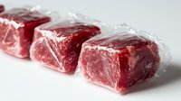Fresh raw beef chunks individually wrapped in clear plastic, arranged in row showing marbling and deep red meat color on white surface for food industry and retail.