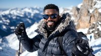 Man with dark skin tone wearing sunglasses and black winter jacket with fur collar holds ski poles on snowy mountain peak with alpine landscape backdrop.