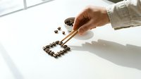 Caucasian hand arranging roasted coffee beans into a square shape using chopsticks on a white surface, with a small bowl nearby.