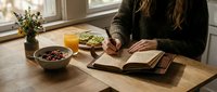 Woman reading book at wooden table with healthy breakfast bowl and orange juice in cozy morning light by window creating peaceful lifestyle moment.