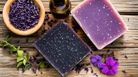 Handmade soap bars with natural lavender flowers and essential oil on rustic wooden background. Purple and charcoal artisanal spa products for skincare.