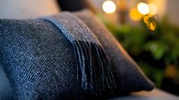 Cozy dark gray wool throw blanket with fringe tassels on sofa, soft bokeh lights and greenery in background creating warm atmospheric home decor setting.