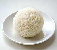 Round ball of steamed white rice served on plain ceramic plate against light background, showing texture and individual grains in natural lighting.