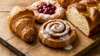 Fresh baked pastries including croissant, cinnamon roll with icing, berry jam tart and sliced bread on rustic wooden cutting board for bakery menu design.