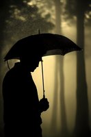 Silhouette of person holding umbrella in rain against misty forest background with golden atmospheric lighting creates moody, contemplative scene.