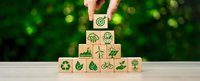 Hand placing wooden cube with target symbol on pyramid of eco-friendly icons representing sustainable business goals and environmental responsibility.
